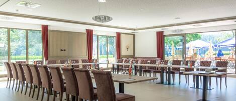 A spacious conference room with multiple tables and chairs, large windows offering a view of the outdoors, and a ceiling with recessed lighting.