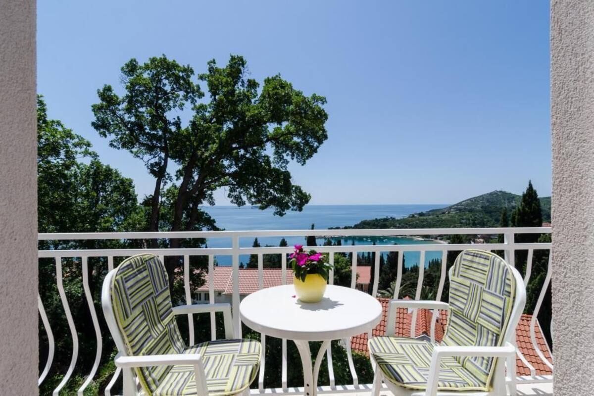 A balcony with two striped chairs and a small round table, overlooking a scenic view of the sea and hills.