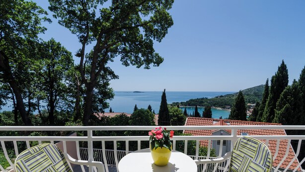 A balcony with a white railing, a round table, and two striped chairs. A yellow vase with pink flowers is on the table. The view includes a body of water and distant buildings. - Apartments Marmo (Zupa dubrovacka)