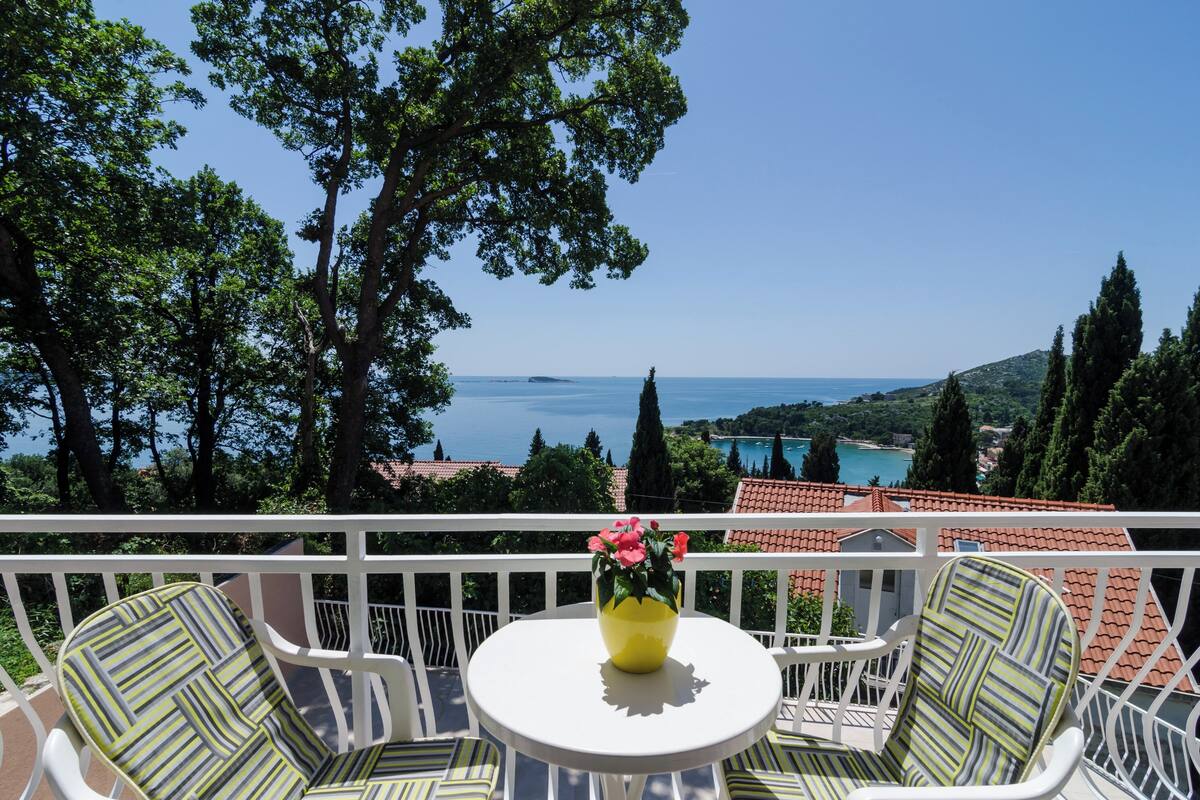 A balcony with a white railing, a round table, and two striped chairs. A yellow vase with pink flowers is on the table. The view includes a body of water and distant buildings.