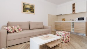 A modern living room with a beige sofa, a white coffee table, and a floral ottoman. A kitchen area with a microwave and cabinets is visible in the background. - Apartments Marmo (Zupa dubrovacka)