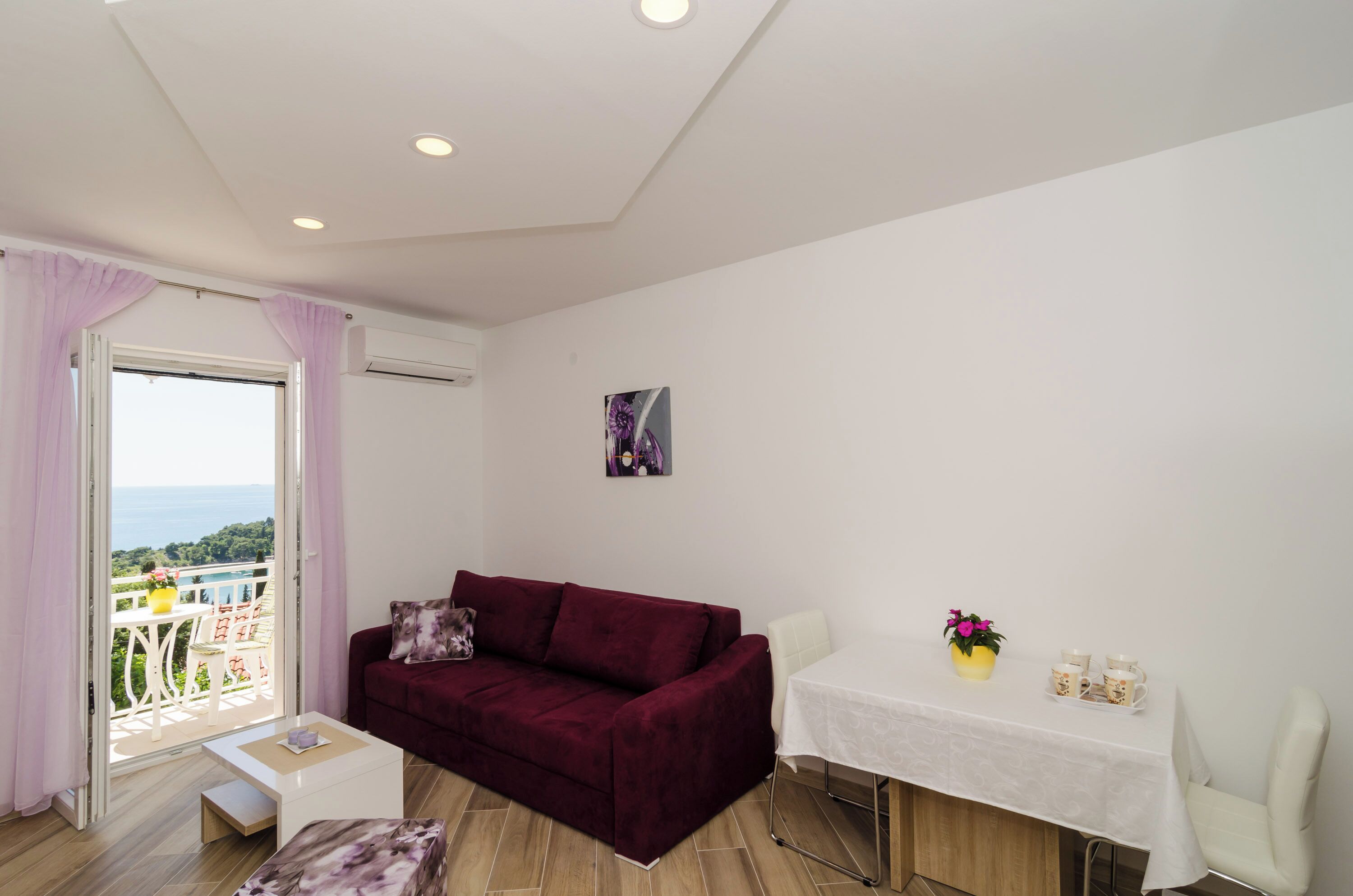 A room with a red sofa, a small white table, a balcony with a view, and a white wall with a picture.