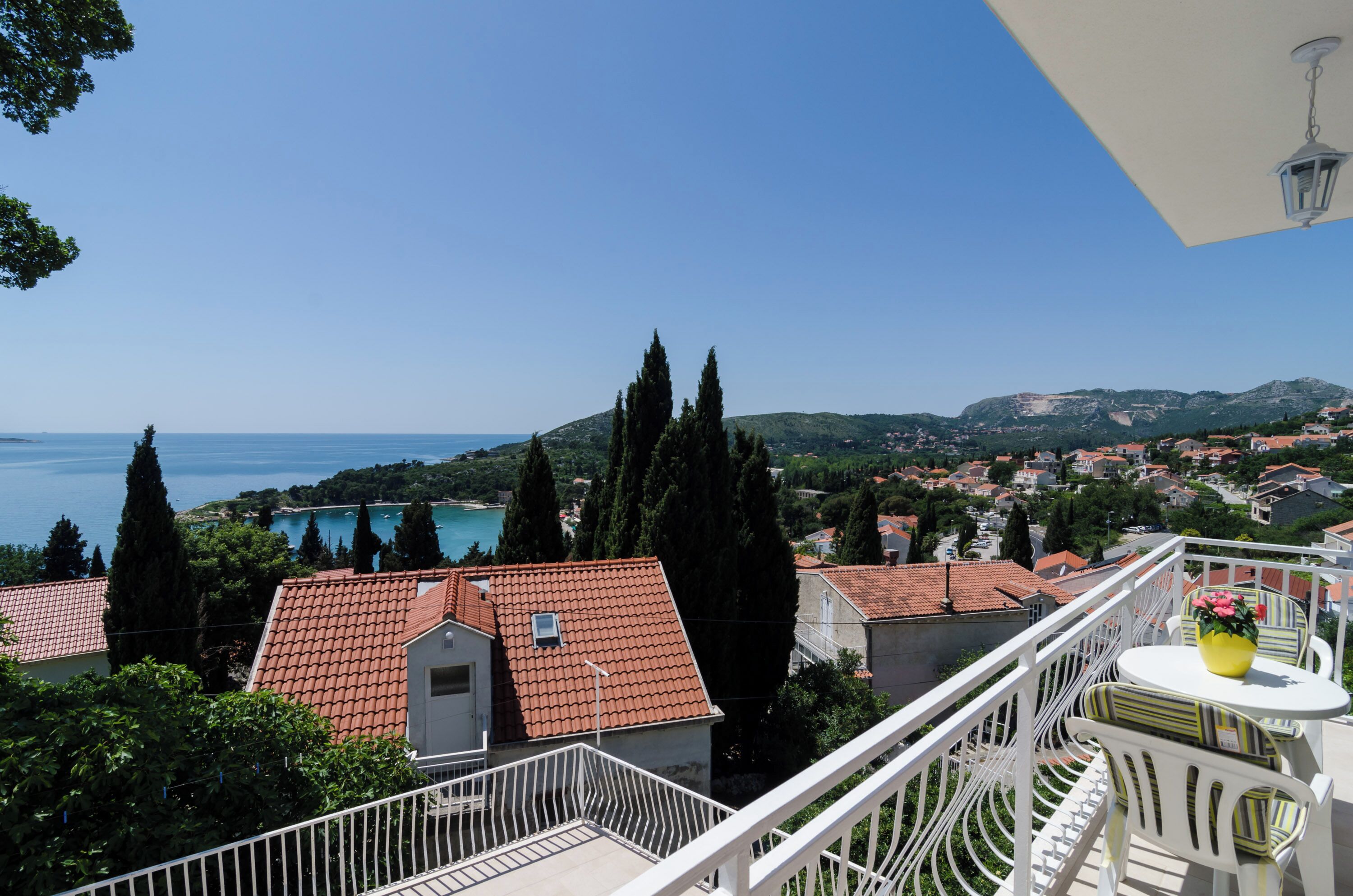 A balcony with white railings overlooking a coastal town with red-roofed buildings and a clear blue sky.