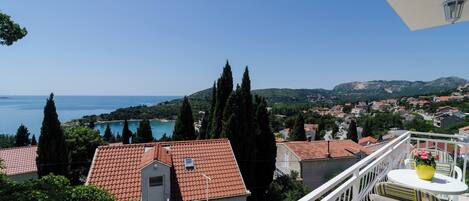 A balcony with white railings overlooking a coastal town with red-roofed buildings and a clear blue sky.