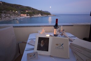 Una mesa dispuesta para una comida, con un menú, una botella de vino y copas, con vistas al mar y la luna en el cielo.