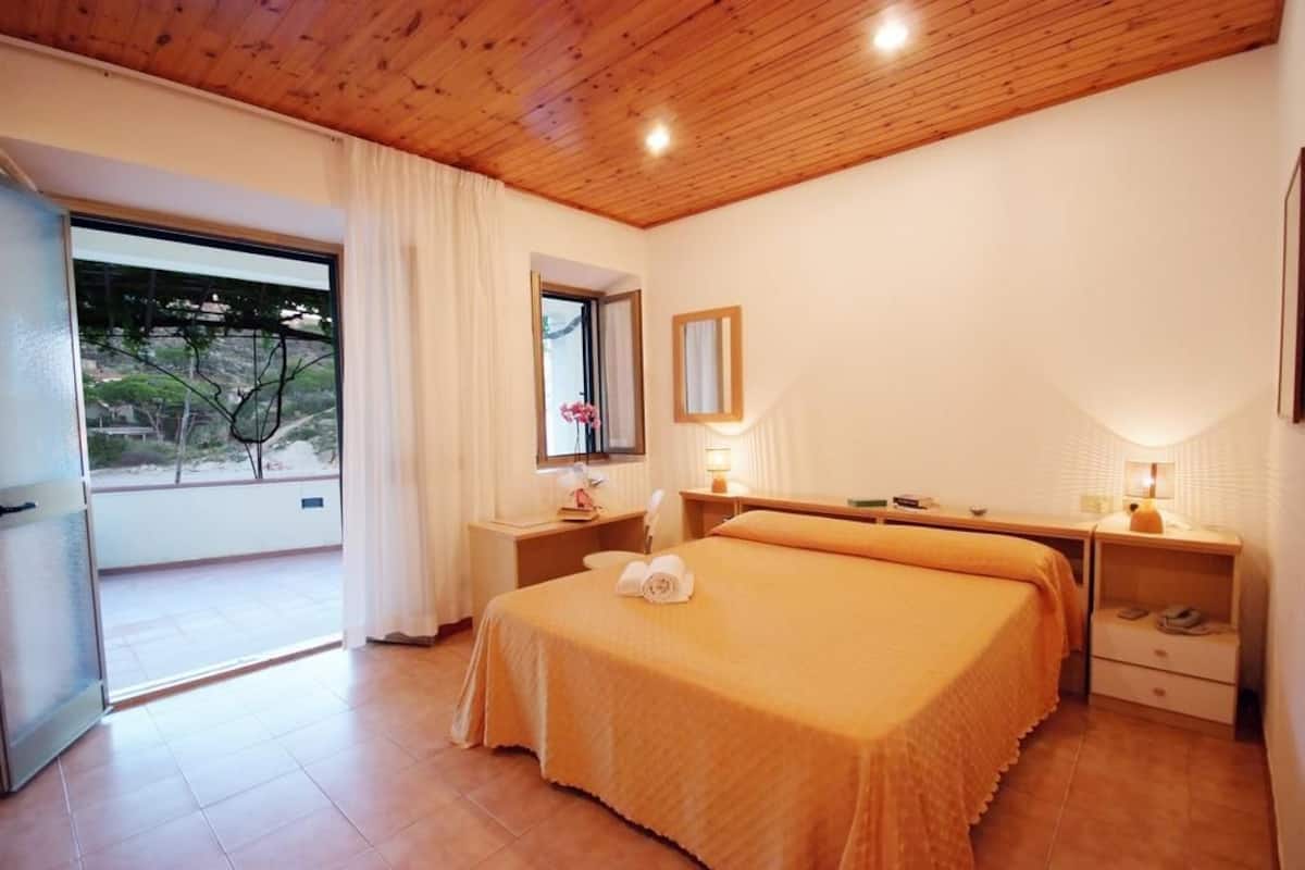 Ein Hotelzimmer mit Holzboden, einem großen Bett mit orangefarbener Bettdecke, einem Fenster mit Blick auf Grünflächen und einem kleinen Tisch mit Lampe.