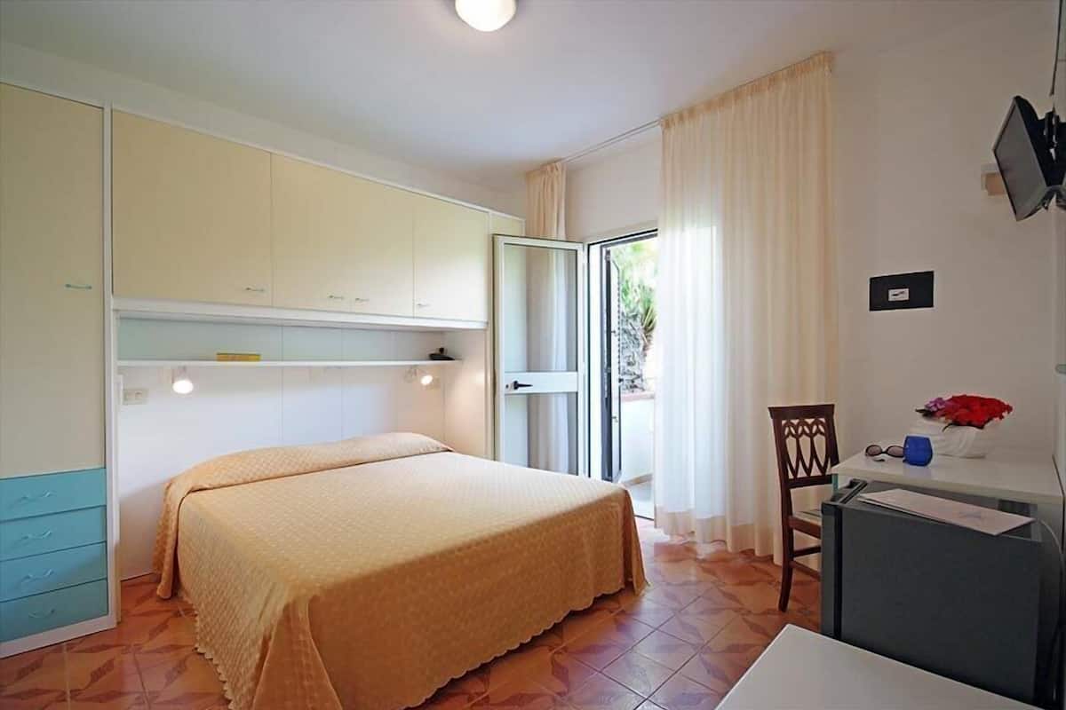 Ein Schlafzimmer mit Bett, Schreibtisch, Stuhl, Kleiderschrank und einem Fenster mit Vorhängen.