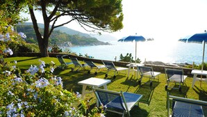 A garden with lawn chairs and tables, a tree, and a view of the sea. - Hotel La Stella (Campo nell'Elba)