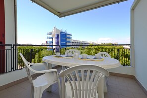 A balcony with a white table and chairs, a view of a blue and white building, and a green fence in the background.