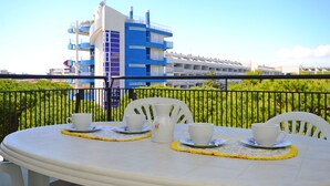 Un balcón con una mesa dispuesta para dos, sillas blancas y una jarrón blanco. Un edificio azul y blanco se ve al fondo.