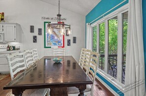 A dining room with a large table, white chairs, a chandelier, a map on the wall, and a window with a view of greenery.
