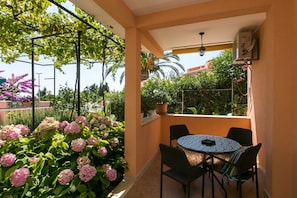 A covered outdoor patio with a round table and chairs, surrounded by lush greenery and flowering plants. - Apartments Bibijana (Zupa dubrovacka)