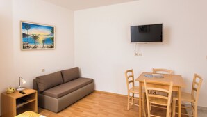 A room with a sofa, a small table with chairs, a TV mounted on the wall, and a painting of a beach scene. - Apartments Bibijana (Zupa dubrovacka)
