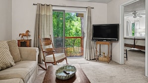 A living room with a sofa, a rocking chair, a television, and a balcony with a view of greenery.