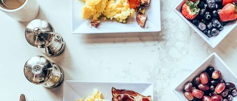 A breakfast setting with plates of scrambled eggs, bacon, and toast, accompanied by bowls of fruit and coffee.