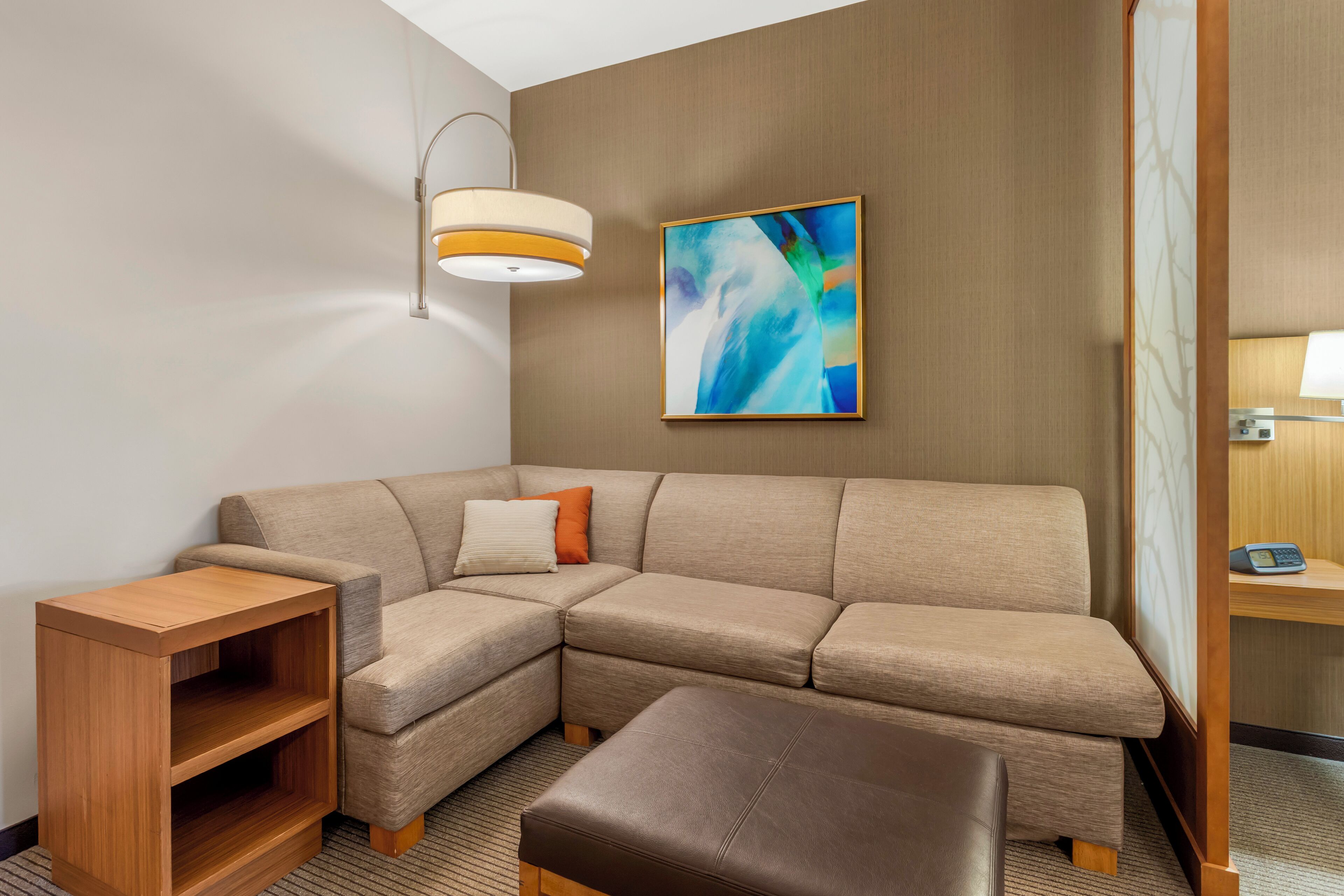 A modern hotel room with a sectional sofa, a wooden side table, a cushioned bench, a wall-mounted abstract painting, and a ceiling light fixture.