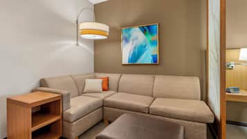 A modern hotel room with a sectional sofa, a wooden side table, a cushioned bench, a wall-mounted abstract painting, and a ceiling light fixture.