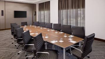 Un salón de conferencias con una larga mesa, varias sillas, un televisor de pantalla plana y ventanas amplias con cortinas.