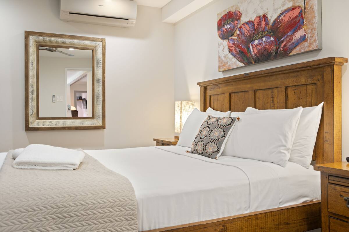 Una cama bien tendida con una colcha blanca y un plaid estampado, un cabecero de madera, una mesita de noche con una lámpara y un espejo grande en la pared.