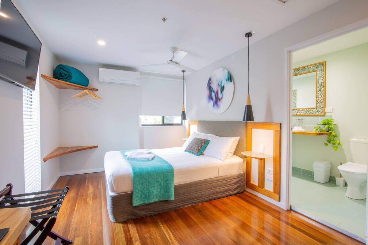 Un dormitorio moderno con cama, piso de madera, ventilador de techo y vista al baño.
