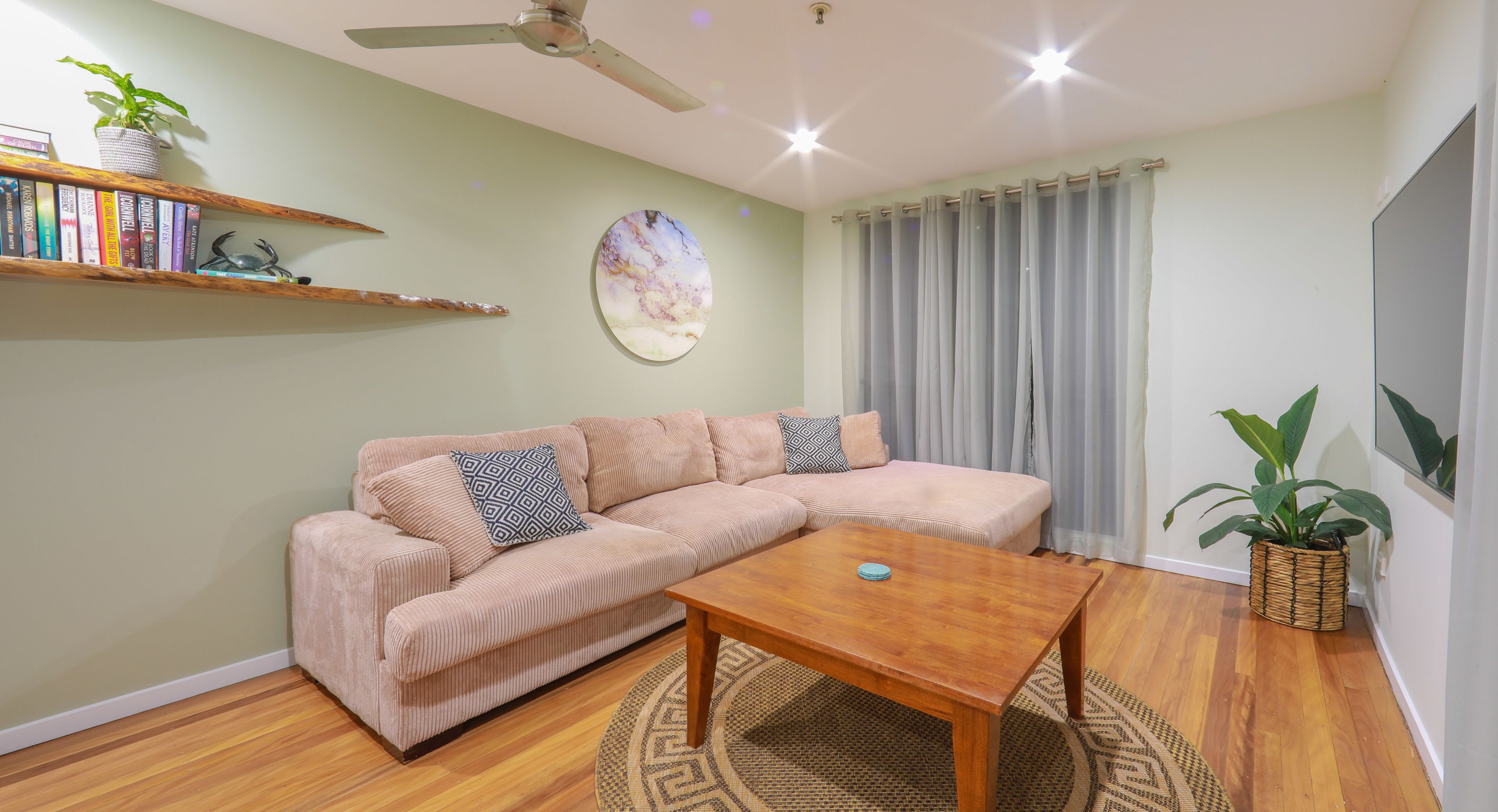 A living room with a beige sofa, a wooden coffee table, a ceiling fan, a wall-mounted shelf with books and a plant, and a large window with curtains.