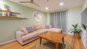 A living room with a beige sofa, a wooden coffee table, a ceiling fan, a wall-mounted shelf with books and a plant, and a large window with curtains.