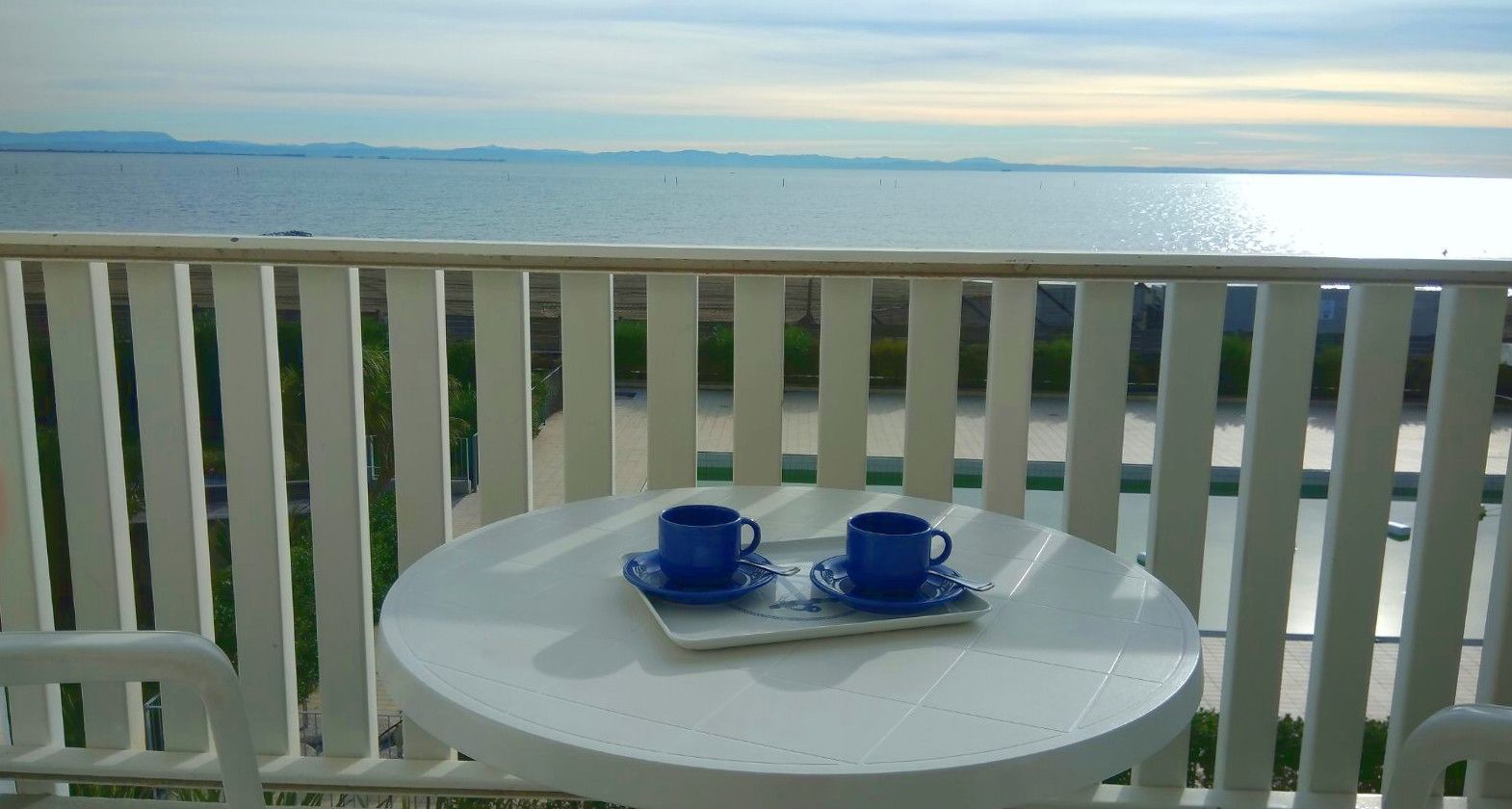 A balcony with a white table and two blue cups on a tray, overlooking a body of water and distant land.