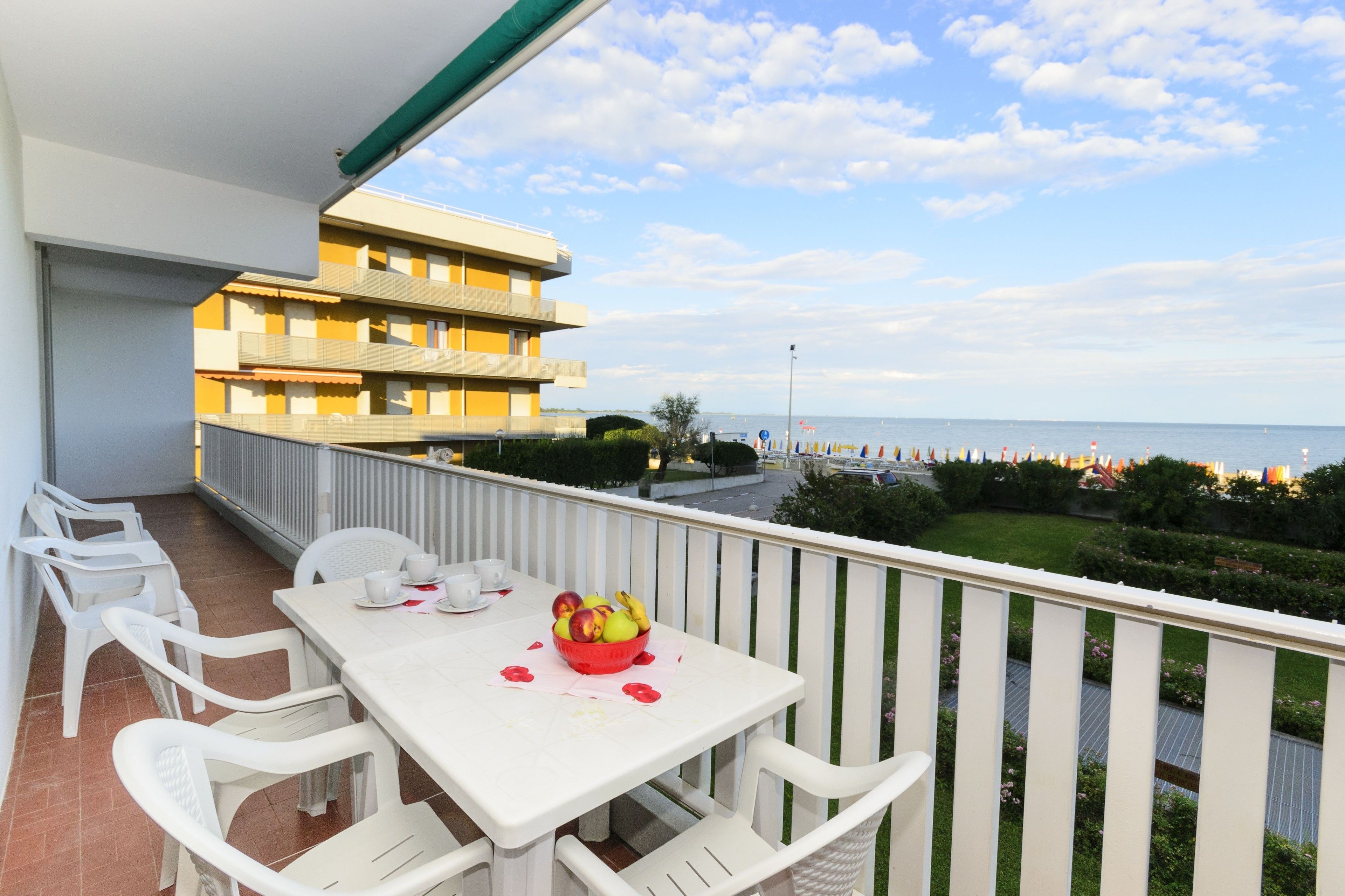 A balcony with a white table and chairs, a bowl of fruit, and a view of the sea and buildings in the background.