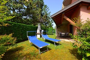 A patio area with two blue lounge chairs, a stone chimney, and a covered seating area with a table and chairs. - Villa Rio (Lignano Sabbiadoro)