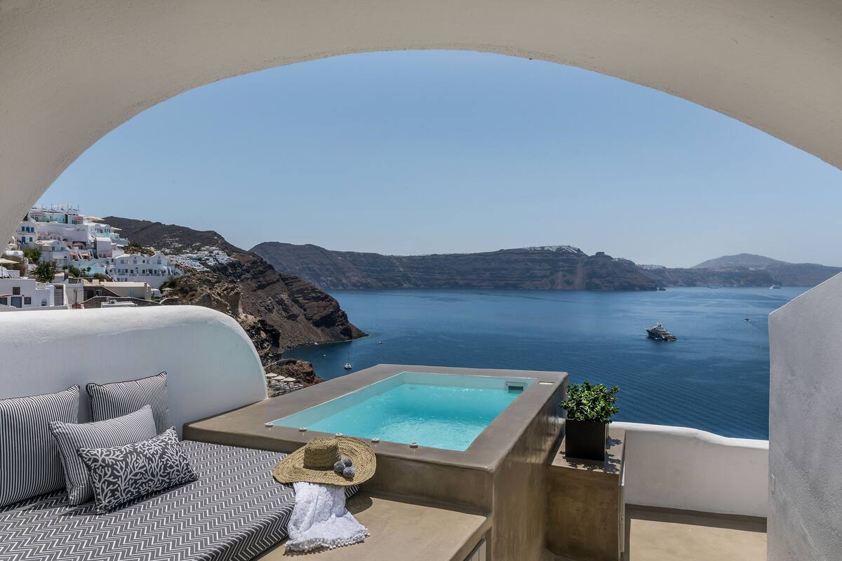 A balcony with a view of the sea, a small pool, and white buildings in the background.