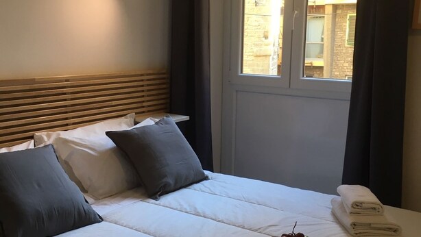 A neatly made bed with white linens, a headboard, and pillows. There is a window with curtains, a wall-mounted light, and a small table with towels and fruit. - Nonna Lina Rooms (Florence)