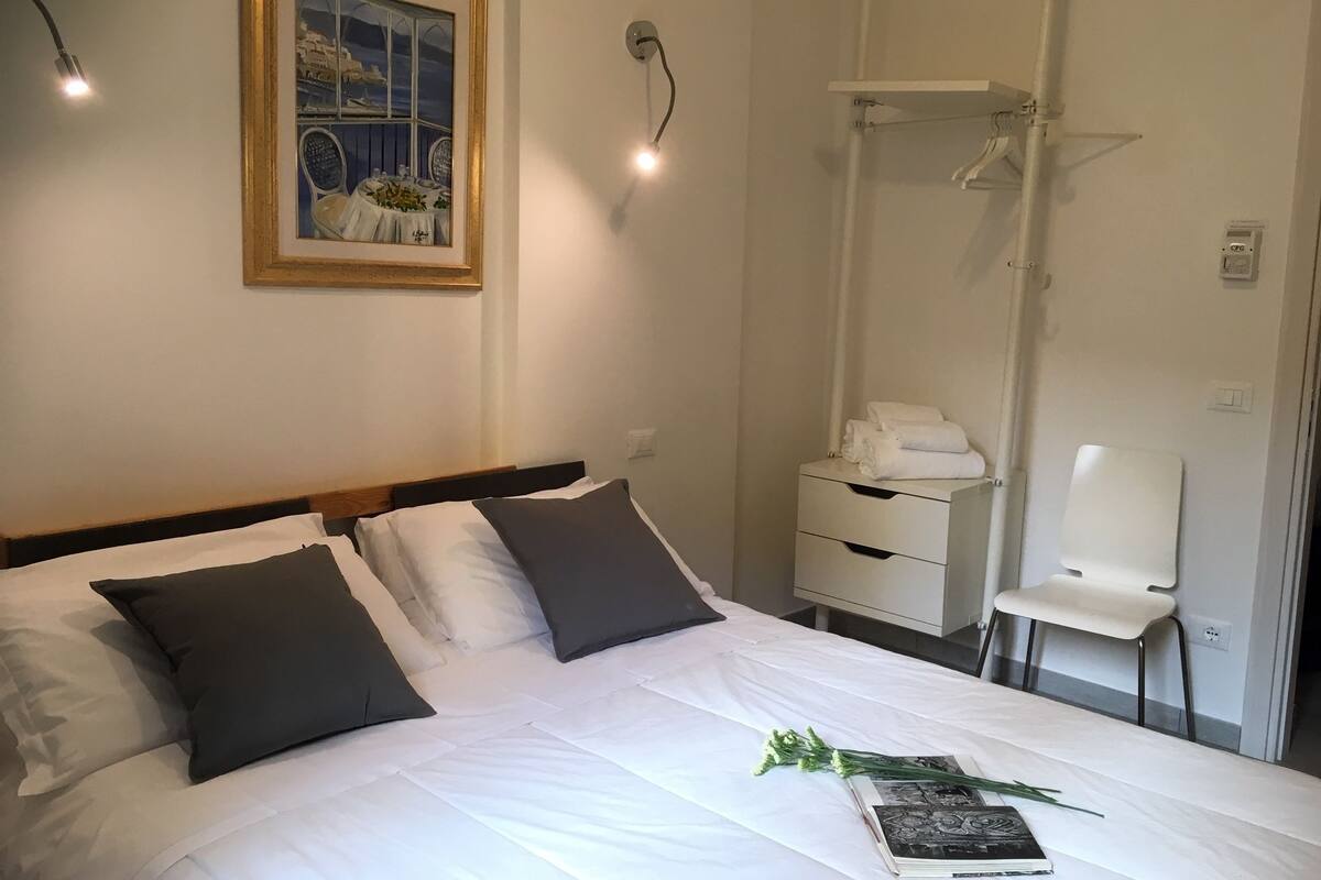 A neatly made bed with white and dark grey pillows, a framed picture on the wall, a bedside table with a lamp, and a chair.