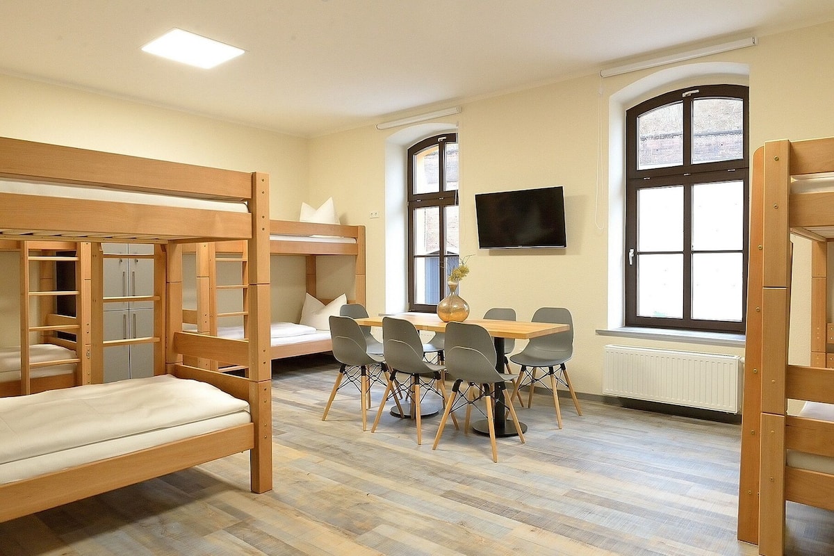 A room with bunk beds, a table with chairs, a flat-screen TV, and large windows.
