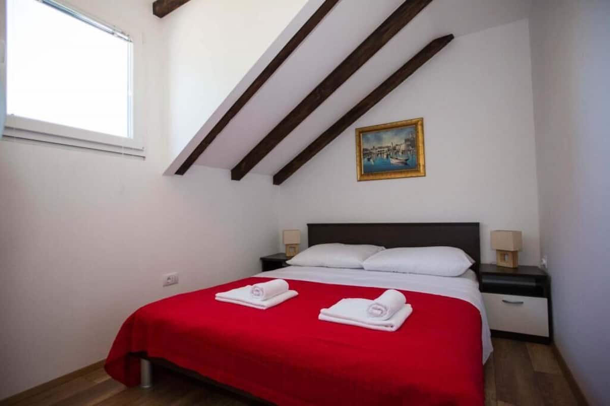 A bedroom with a red bedspread, a dark headboard, two white pillows, a nightstand with a lamp, and a painting on the wall.