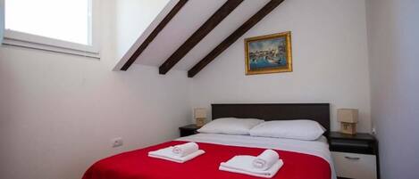Una camera da letto con una coperta rossa, una testata scura, due cuscini bianchi, un comodino con una lampada e un quadro appeso al muro.