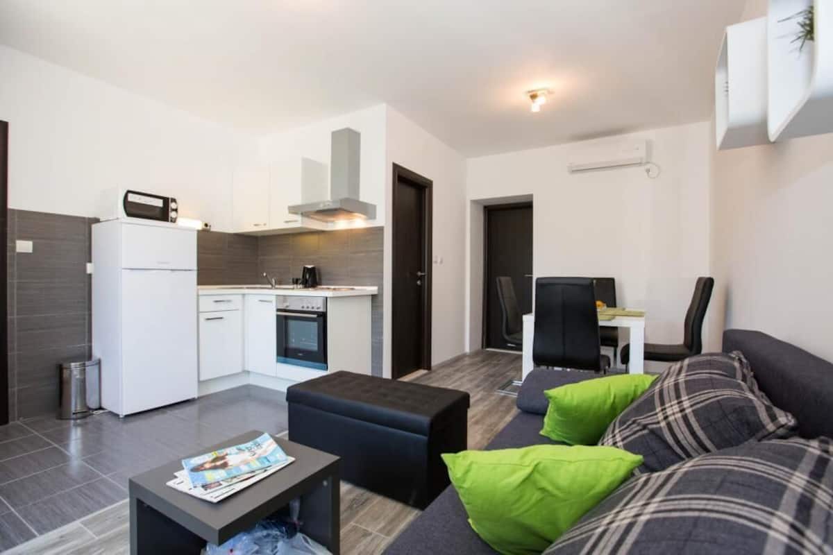 A compact living space with a kitchenette, a sofa with green and plaid cushions, and a small table with a magazine.