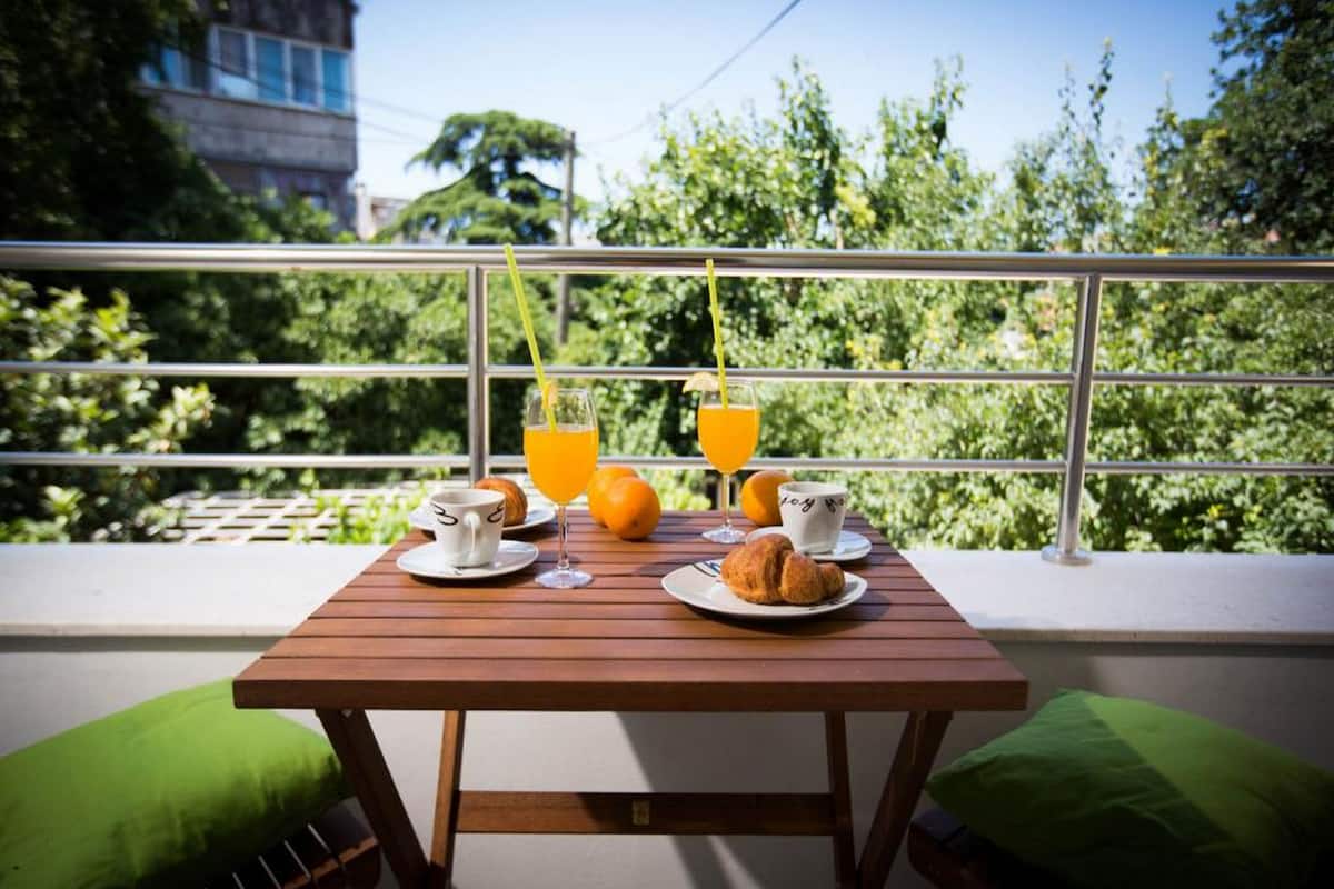 A balcony with a wooden table set for breakfast, including croissants, orange juice, and a cup.