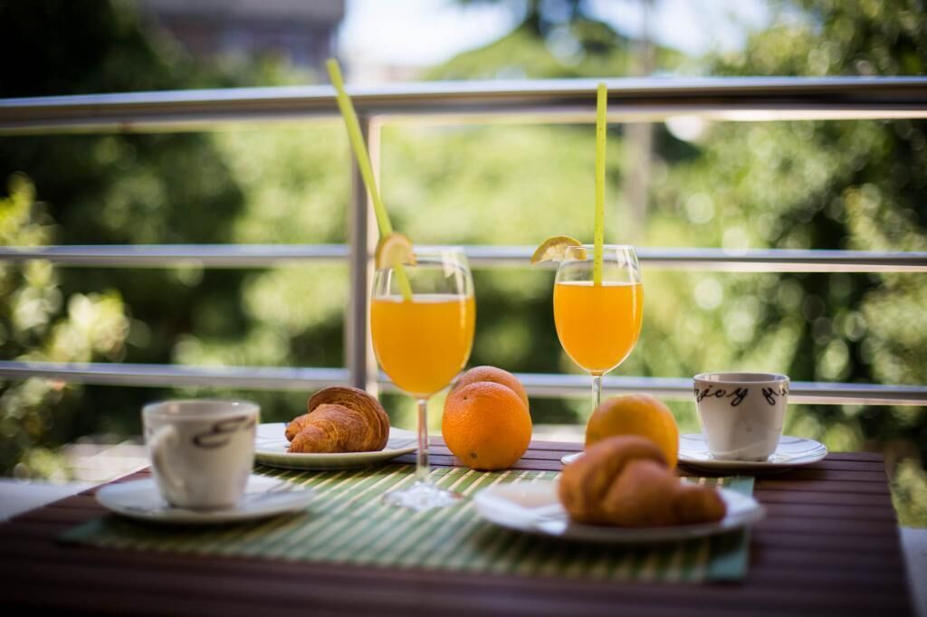A breakfast setting with two glasses of orange juice, a croissant, an orange, and a cup on a table with a view of greenery through a window.