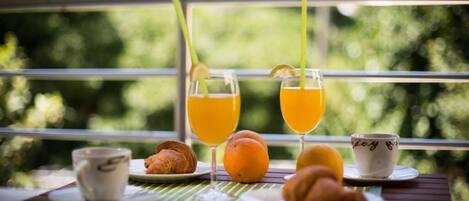 A breakfast setting with two glasses of orange juice, a croissant, an orange, and a cup on a table with a view of greenery through a window.