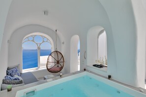 A modern indoor pool area with a view of the sea, a hanging chair, and a seating area with patterned cushions. - Namaste Suites by Oias Local Cavehouses (Santorini)