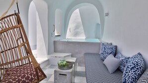 Een moderne kamer met een rieten stoel, een bank met decoratieve kussens, een klein tafeltje met een fruitschaal, en een badkuip met uitzicht op de lucht.