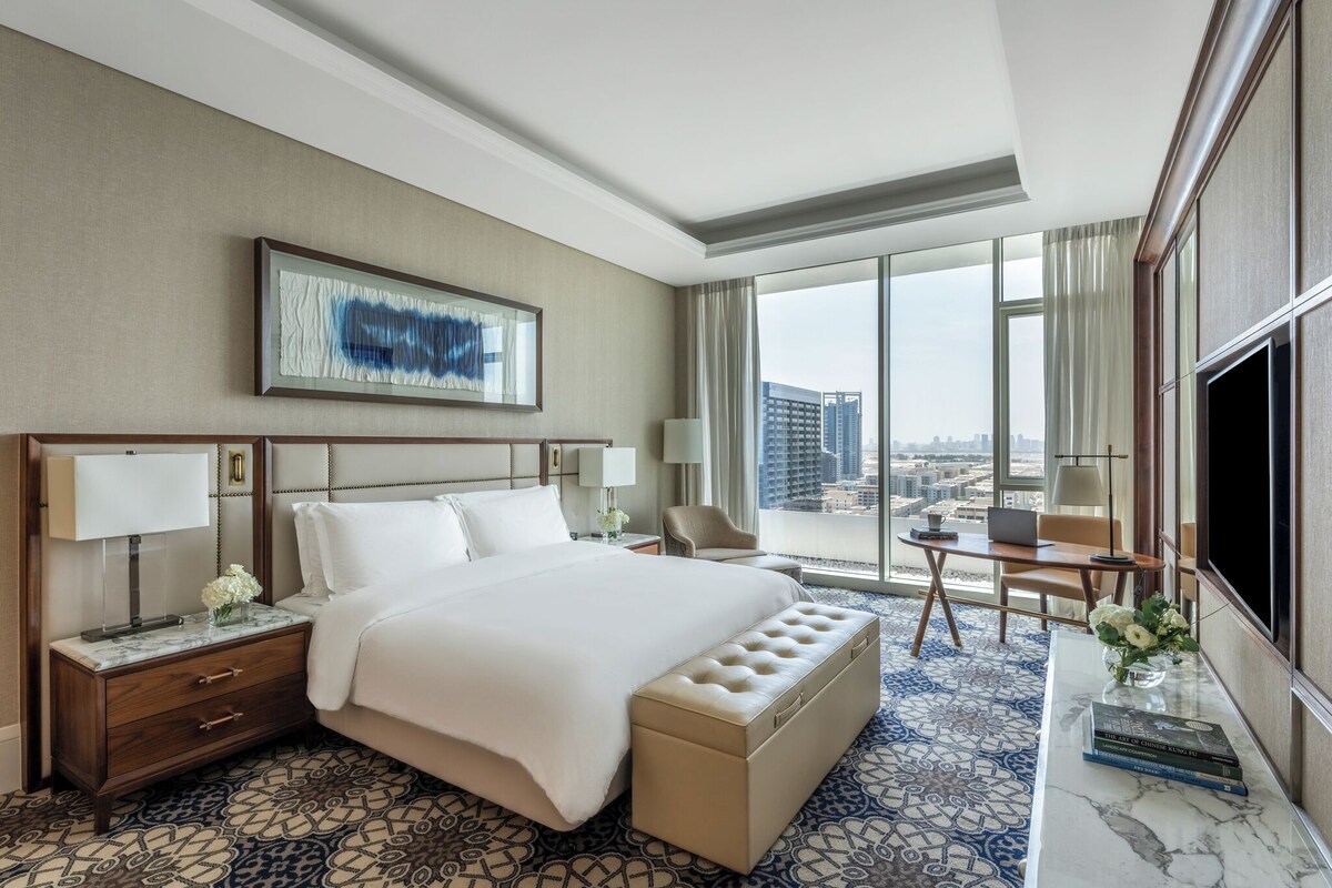 A hotel room with a large bed, a desk with a laptop, a TV, and a view of the city.