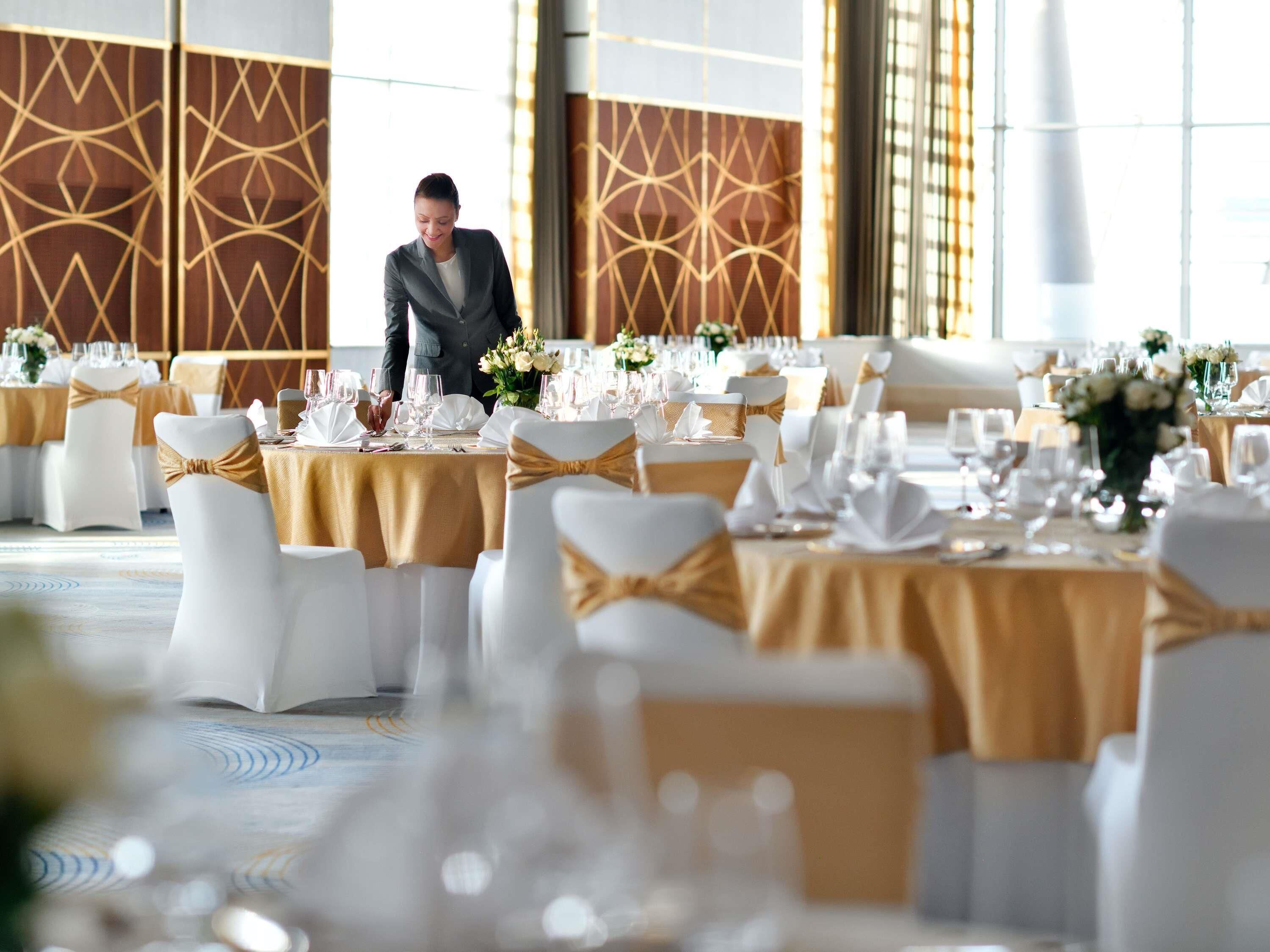 A banquet hall with round tables set for dining, white chairs with gold sashes, and a person in a suit standing in the background.