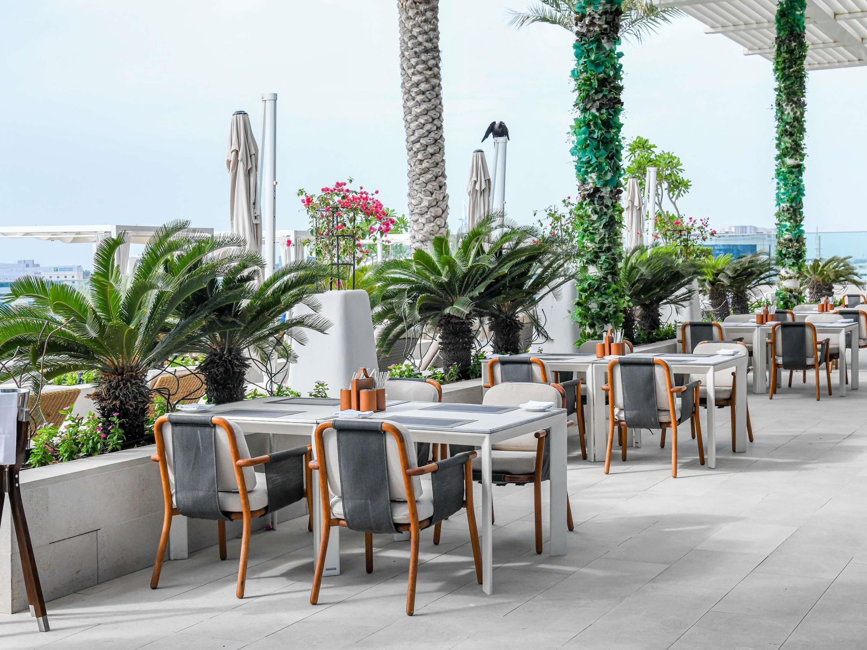 An outdoor dining area with tables and chairs, surrounded by potted plants and umbrellas.