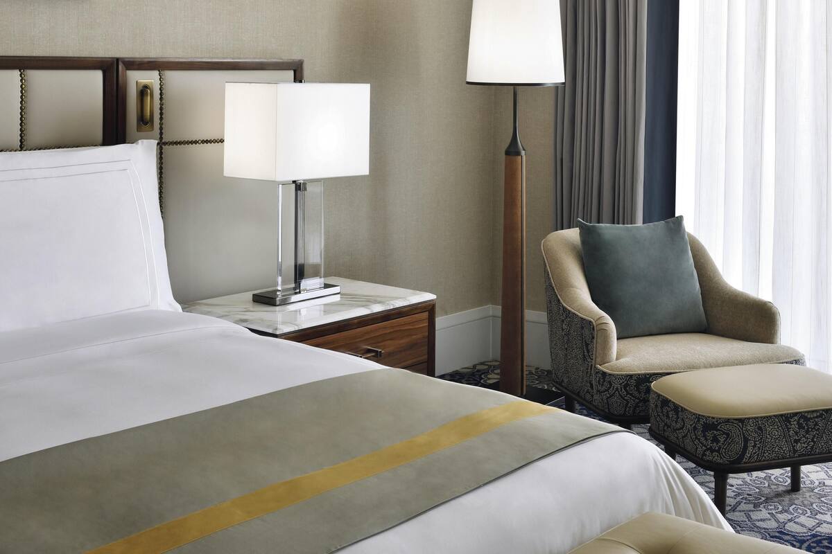 Una habitación de hotel con cama, mesitas de noche, lámpara, una silla y ventana con cortinas.