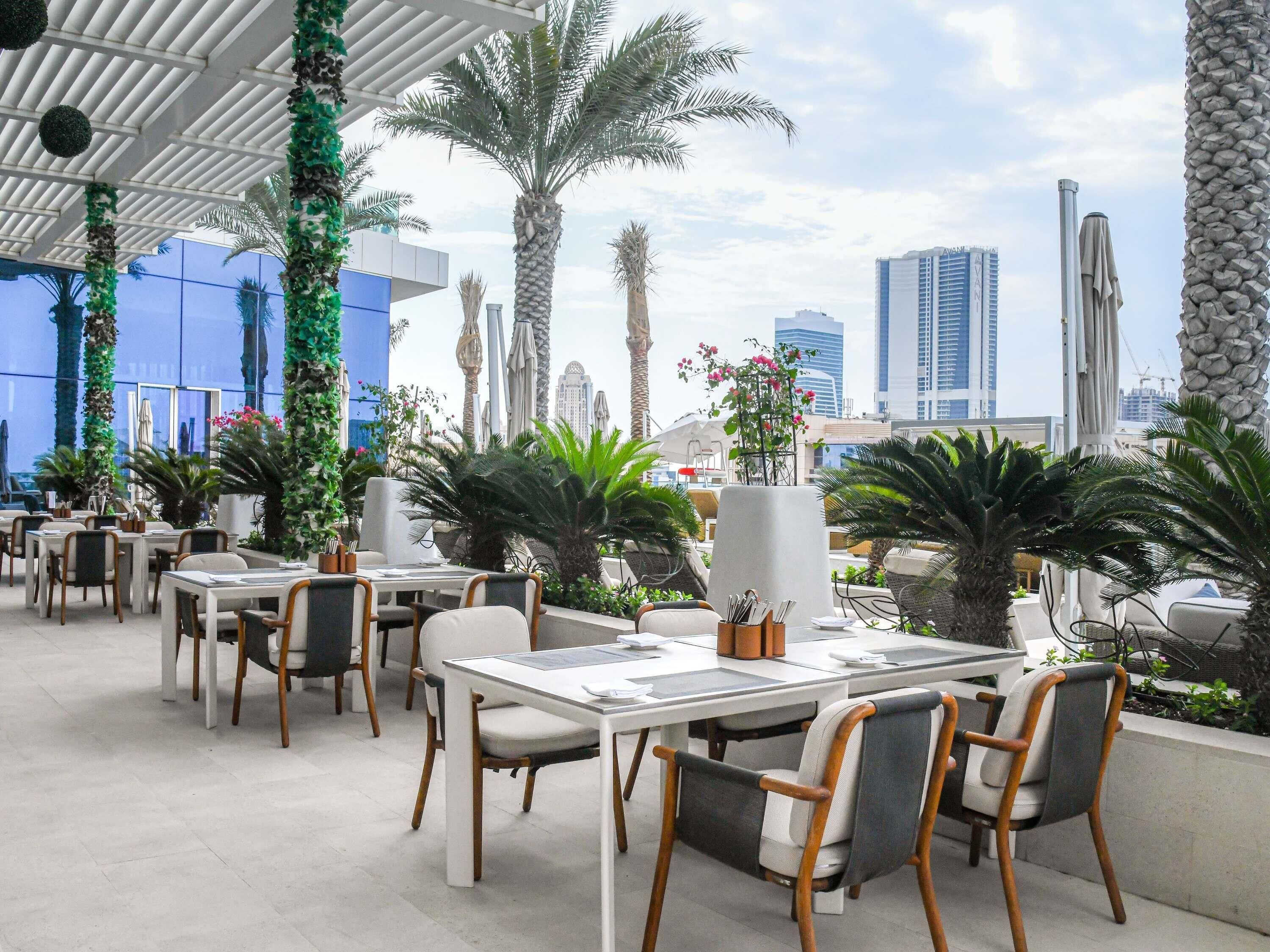 Un espace de restauration en plein air, avec des tables et des chaises, entouré de verdure et de palmiers, offrant une vue sur un paysage urbain avec des bâtiments modernes.