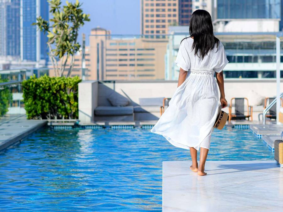 Uma mulher de vestido branco caminhando perto de uma piscina, com paisagem urbana ao fundo.