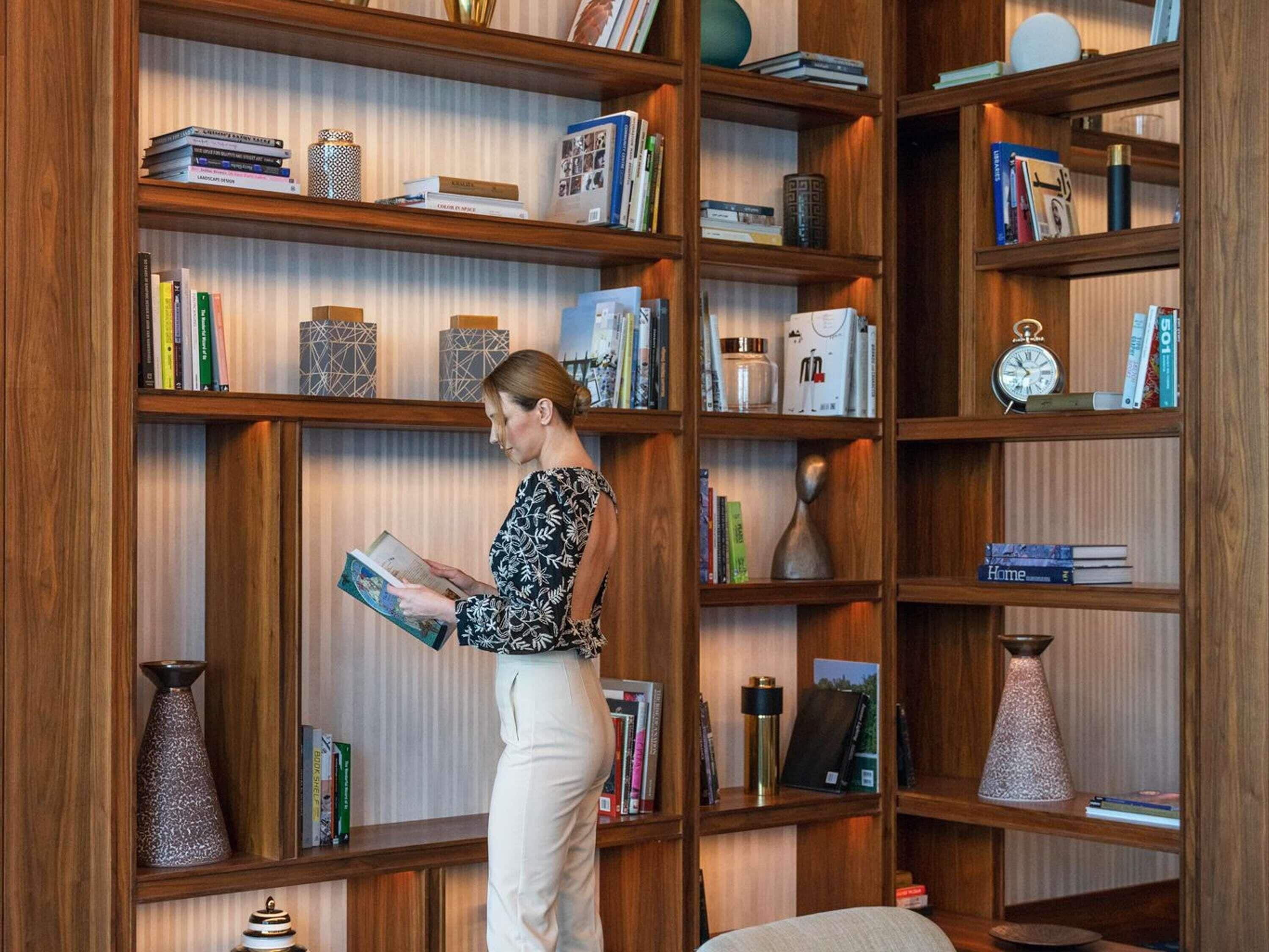 Eine Frau liest ein Buch in einem Raum mit Holzregalen, die mit Büchern und Dekorationsgegenständen gefüllt sind.