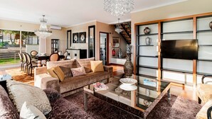 A spacious living room with a large sofa, a coffee table, and a TV mounted on a wall. There is a staircase leading to an upper level, and a dining area with chairs and a table.