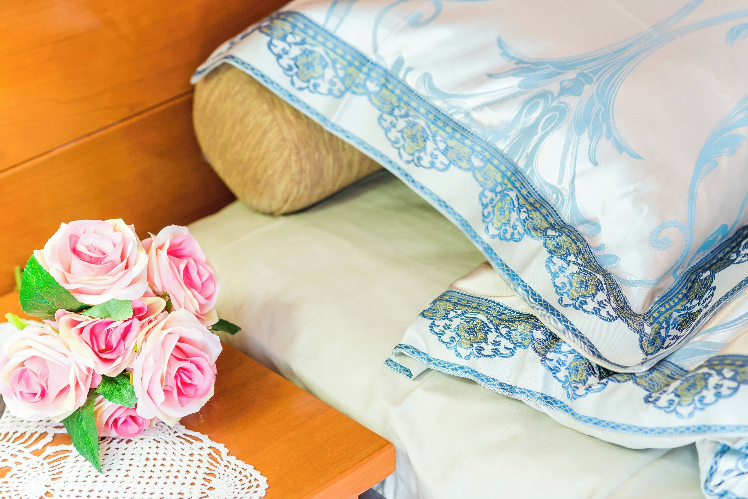 A bed with a blue and white patterned blanket, a wooden headboard, and a bouquet of pink roses on a wooden nightstand.